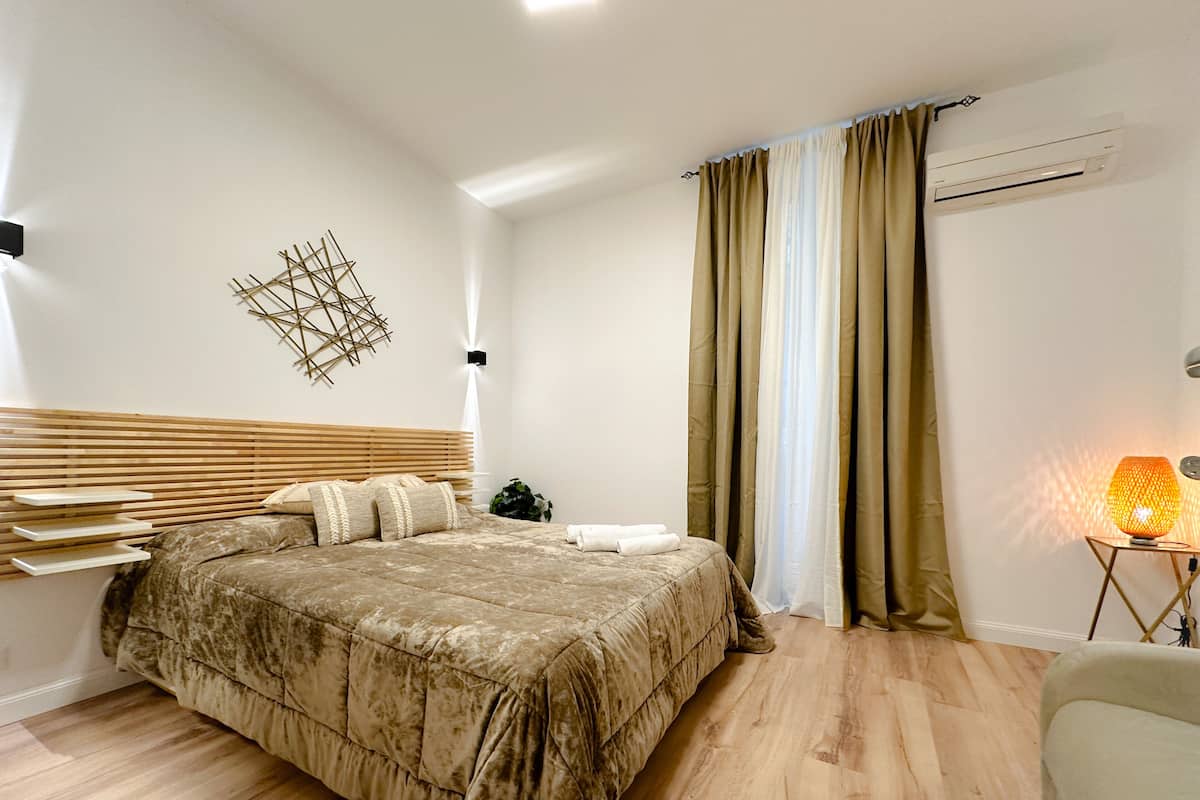A bedroom with a large bed, a bedside table, a wall-mounted air conditioner, and a floor lamp.