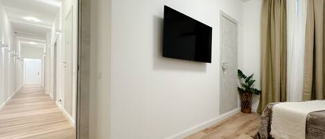 Habitación de hotel con piso de madera, cama con frazada color crema, televisor de pantalla plana en la pared, planta en maceta y puerta que da a otra habitación.
