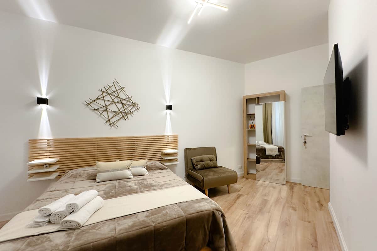 A modern bedroom with a wooden headboard, a bed with rolled towels, a chair, a wardrobe, and a TV mounted on the wall.