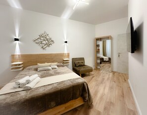 Un dormitorio moderno con cabecera de madera, cama con toallas enrolladas, una silla, un armario y un televisor montado en la pared.