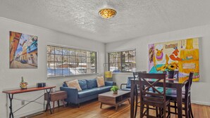 Een woonkamer met een blauwe bank, een houten salontafel en een eethoek met een houten tafel en stoelen. Er zijn twee ramen met metalen tralies, en aan de muur hangt een schilderij.