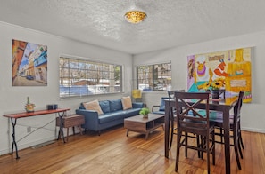 A living room with a blue sofa, wooden coffee table, and a dining area with a wooden table and chairs. There are two windows with metal bars, and a painting is on the wall.