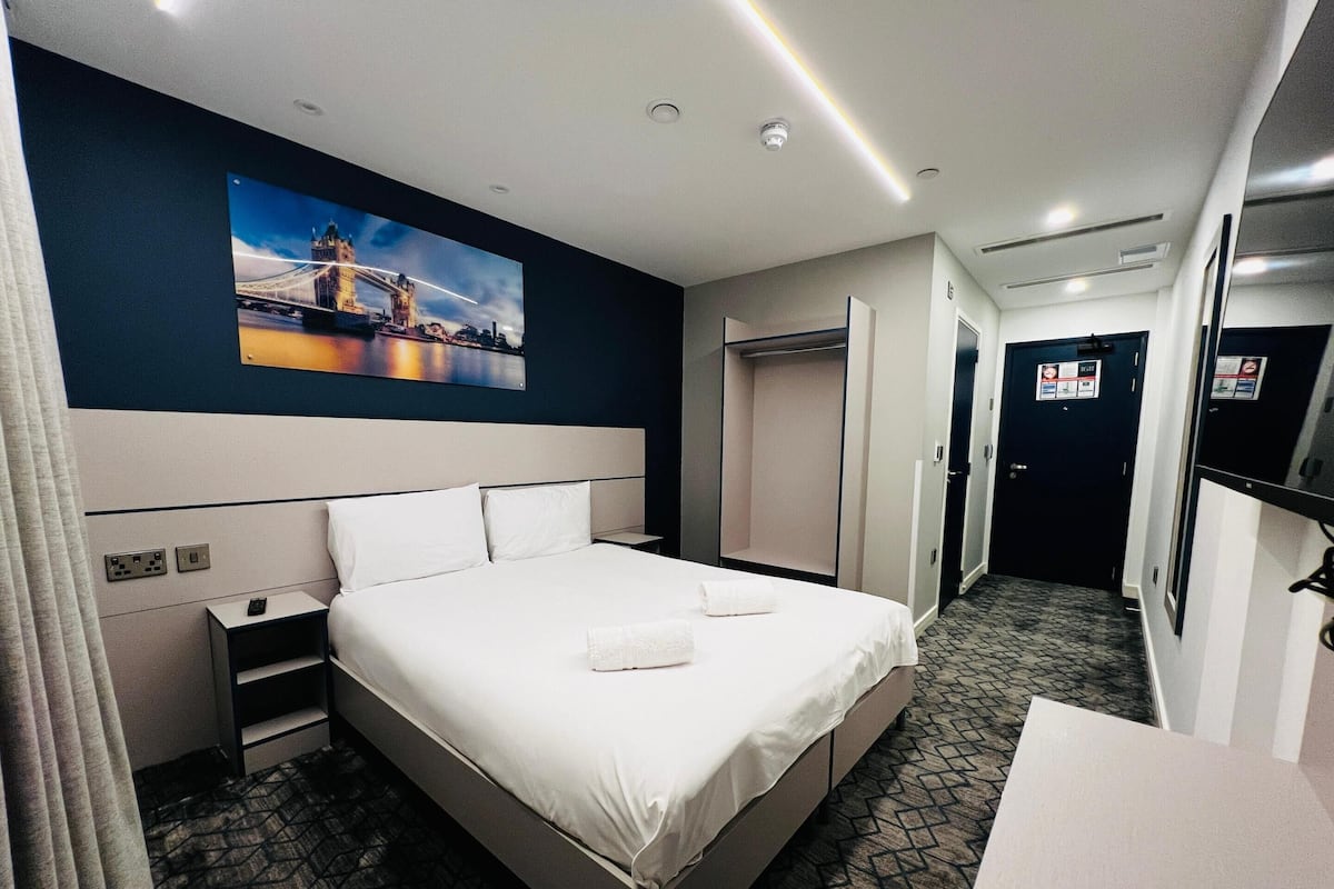 A hotel room with a large bed, a nightstand, a desk, a mirror, and a wardrobe.
