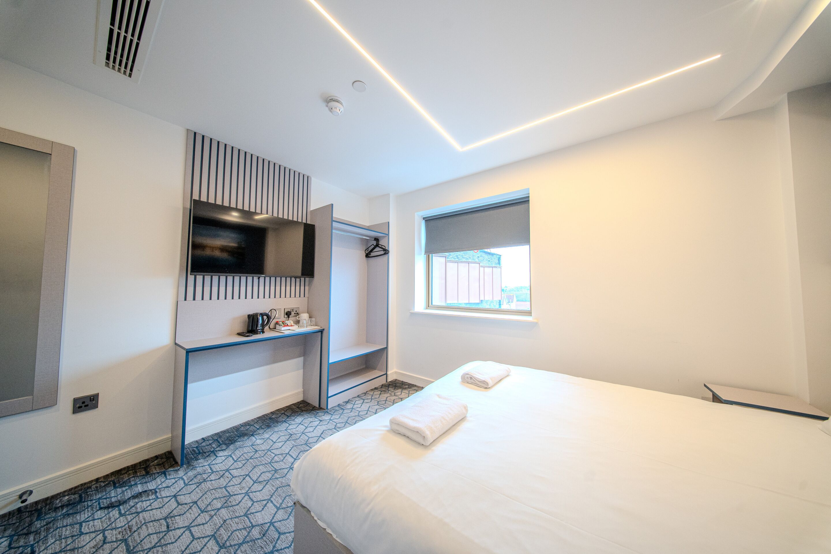 Une chambre d’hôtel moderne équipée d’un lit, d’un téléviseur fixé au mur, d’un bureau avec une chaise et d’une fenêtre laissant entrer la lumière naturelle.