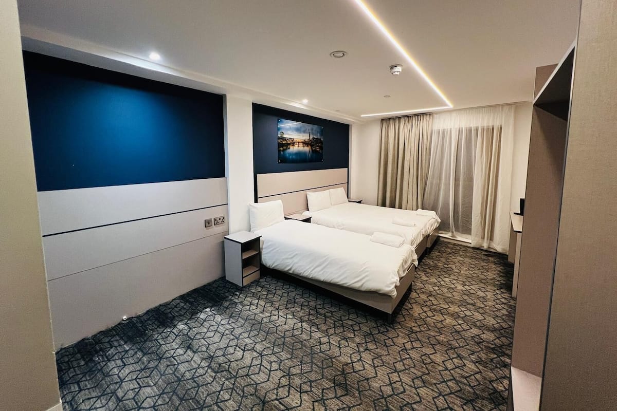 A hotel room with two beds, a large window with curtains, a painting on the wall, and a patterned carpet.