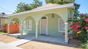 Une maison de plain-pied à la façade vert clair, dotée d’un porche avec trois arcades et d’un petit jardin agrémenté de fleurs roses.