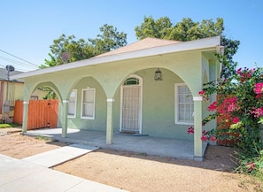 A single-story house with a light green exterior, a front porch with three arches, and a small garden with pink flowers.