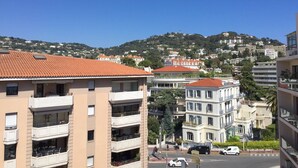 Ein mehrstöckiges Gebäude mit Balkonen, einem roten Ziegeldach und Blick auf eine hügelige Stadtlandschaft im Hintergrund.
