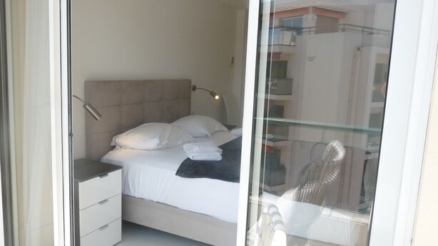 Ein Schlafzimmer mit Bett, Nachttisch, Lampe und Blick durch eine Glastür nach draußen.