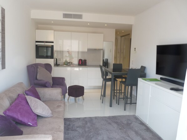 Una sala de estar moderna con un sofá, una mesa de centro y un televisor. Se observa una zona de cocina con armarios y una mesa de comedor.