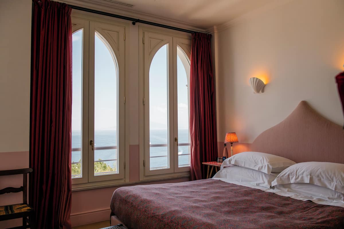 A hotel room with a large bed, a nightstand with a lamp, and a view of the sea through arched windows.