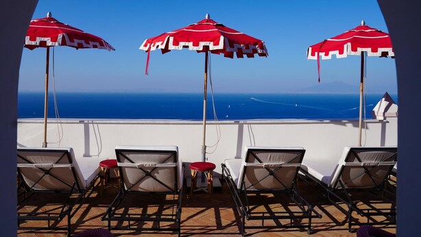 Una terraza con tres sombrillas rojas y blancas, sillones blancos para lounge y vista al océano.