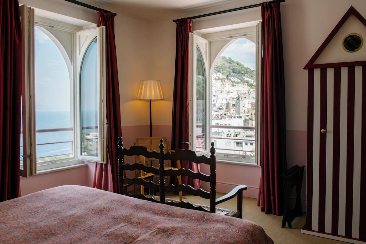 A bedroom with a bed, a chair, a lamp, and a wardrobe. There are two windows with curtains, and a view of a cityscape.