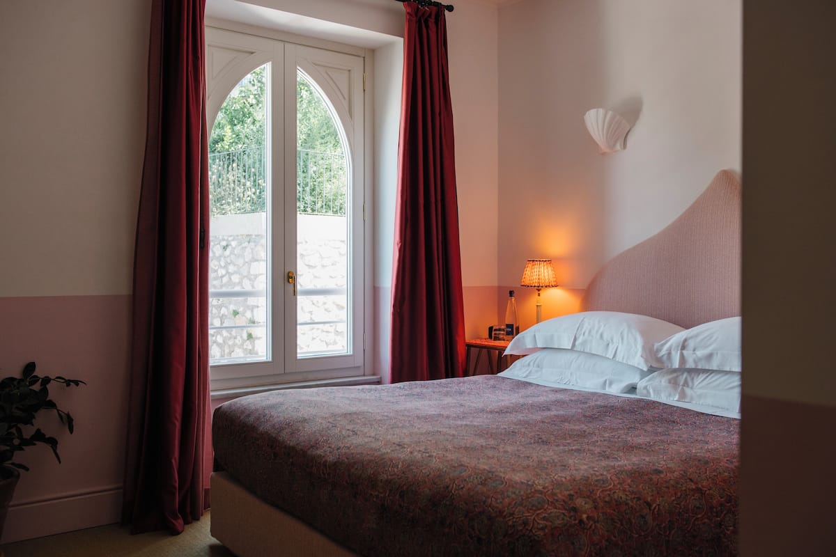 A hotel room with a bed, a nightstand, a lamp, and a view of a stone wall and greenery outside.