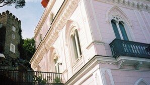 Un edificio rosa con ventanas arqueadas y balcones, una torre de piedra y sombrillas rojas.
