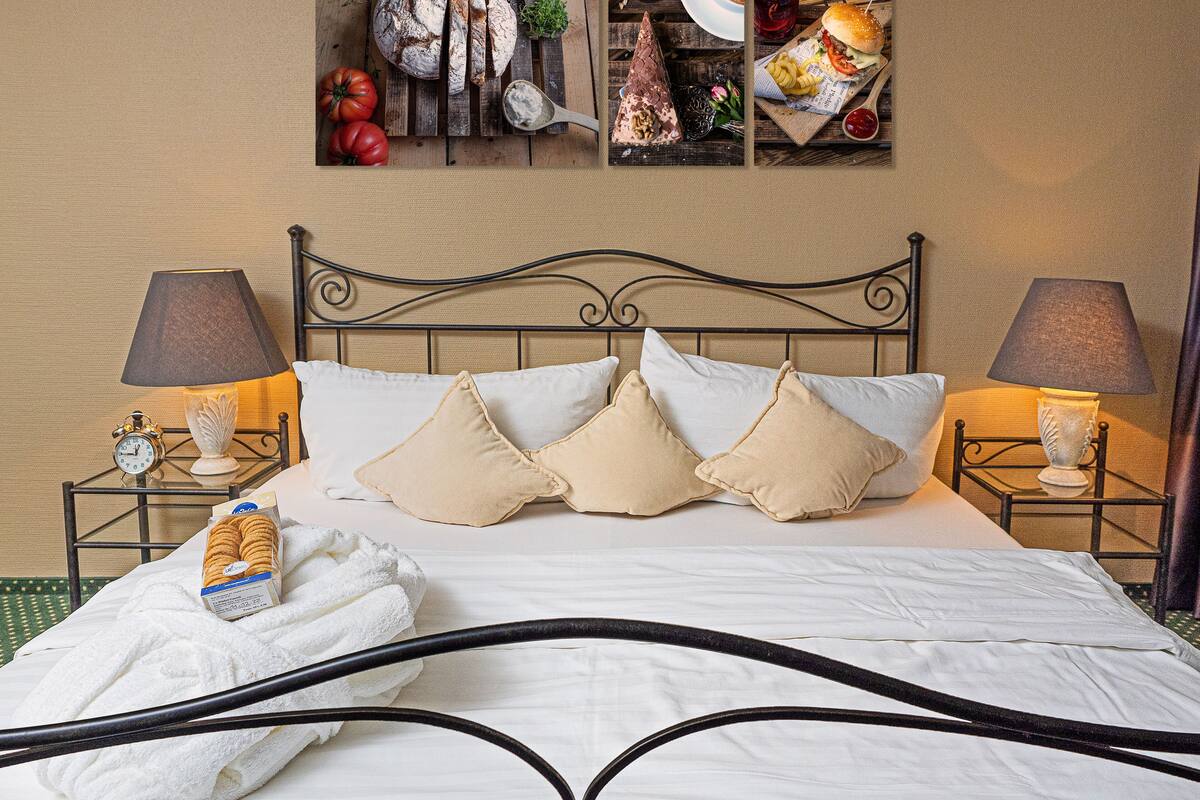 A neatly made bed with white linens and beige pillows, a bedside table with a lamp, and a wall-mounted picture of food items.