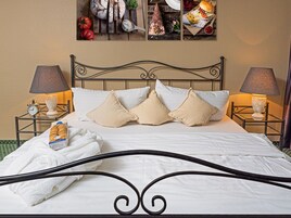 Una cama bien hecha con sábanas blancas y almohadas color beige, una mesita de noche con una lámpara, y un cuadro en la pared con imágenes de comida.