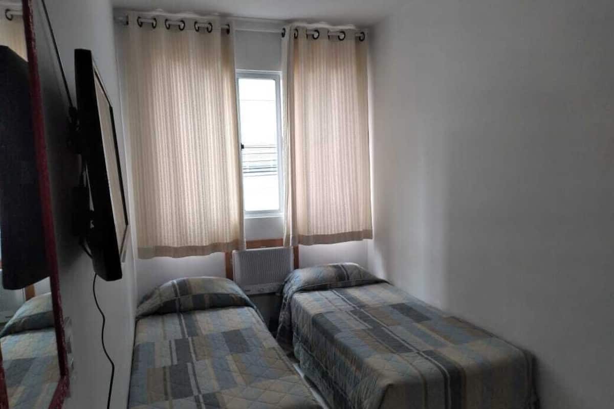 Una habitación pequeña con dos camas individuales, una ventana con cortinas y un televisor montado en la pared.