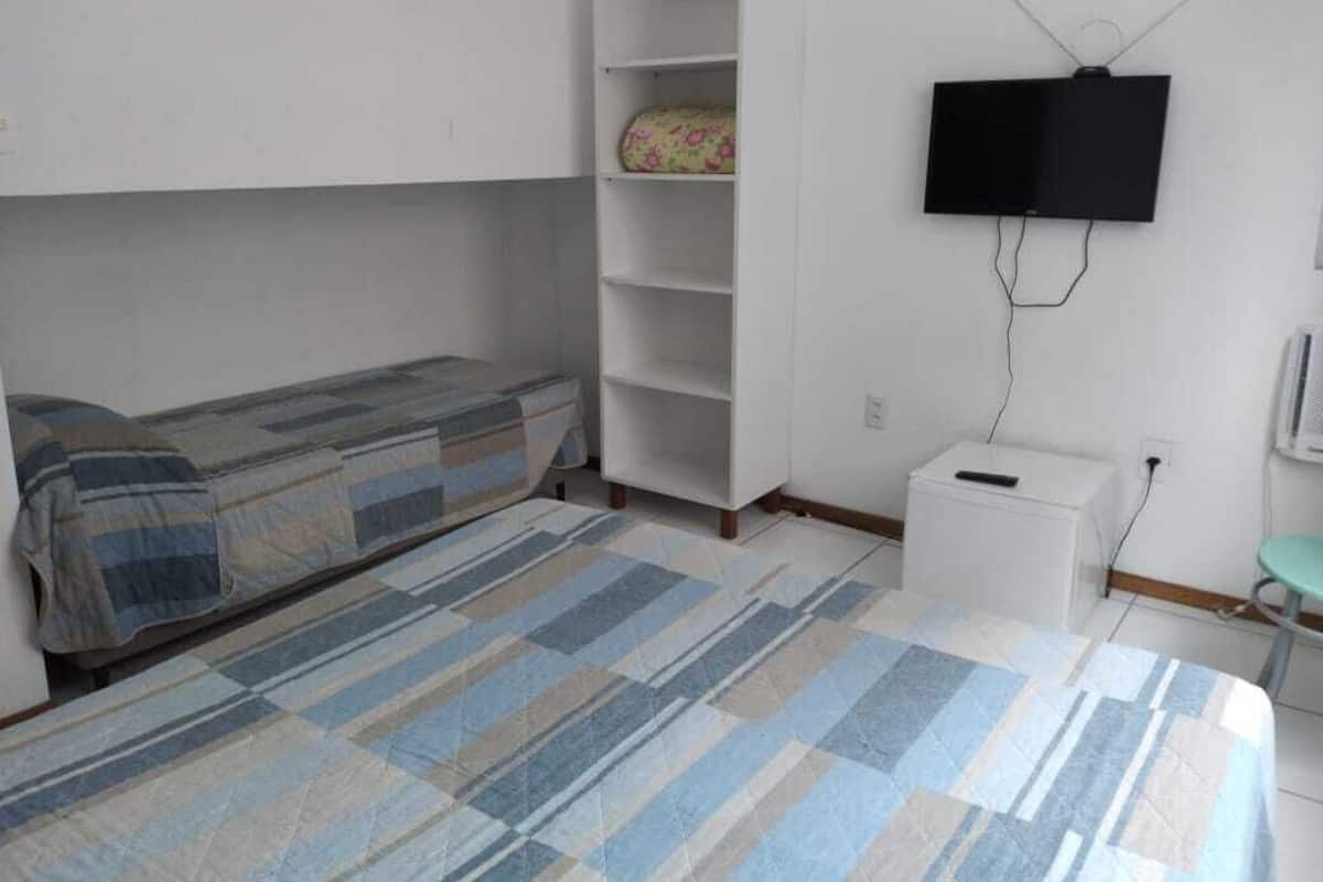 Una habitación pequeña y poco amueblada, con cama, un armario blanco, televisor fijado a la pared y una mesita blanca.