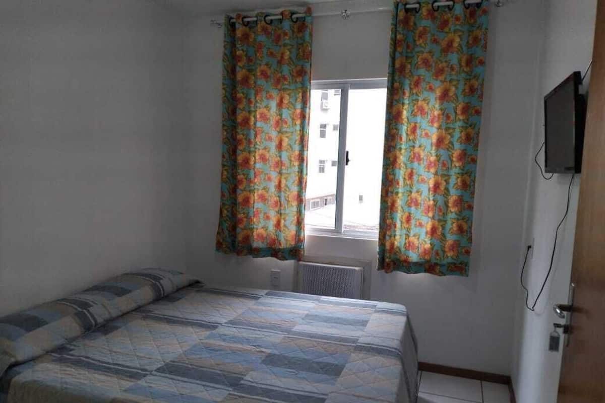 Un dormitorio con cama, cortinas, ventana, radiador y televisor montado en la pared.
