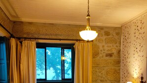 A cozy room with a bed, a bench, and a chair. There is a window with curtains, a lamp, and a stone wall. - Lugar da Pedra Alta (Gouveia)
