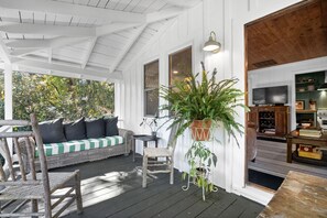 Une véranda abritée avec un canapé en osier, une chaise en bois et une plante suspendue.