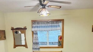 Ein Essbereich mit einem Holztisch, vier Stühlen, einem Deckenventilator, einem Fenster mit gemusterten Vorhängen und einem Spiegel an der Wand.