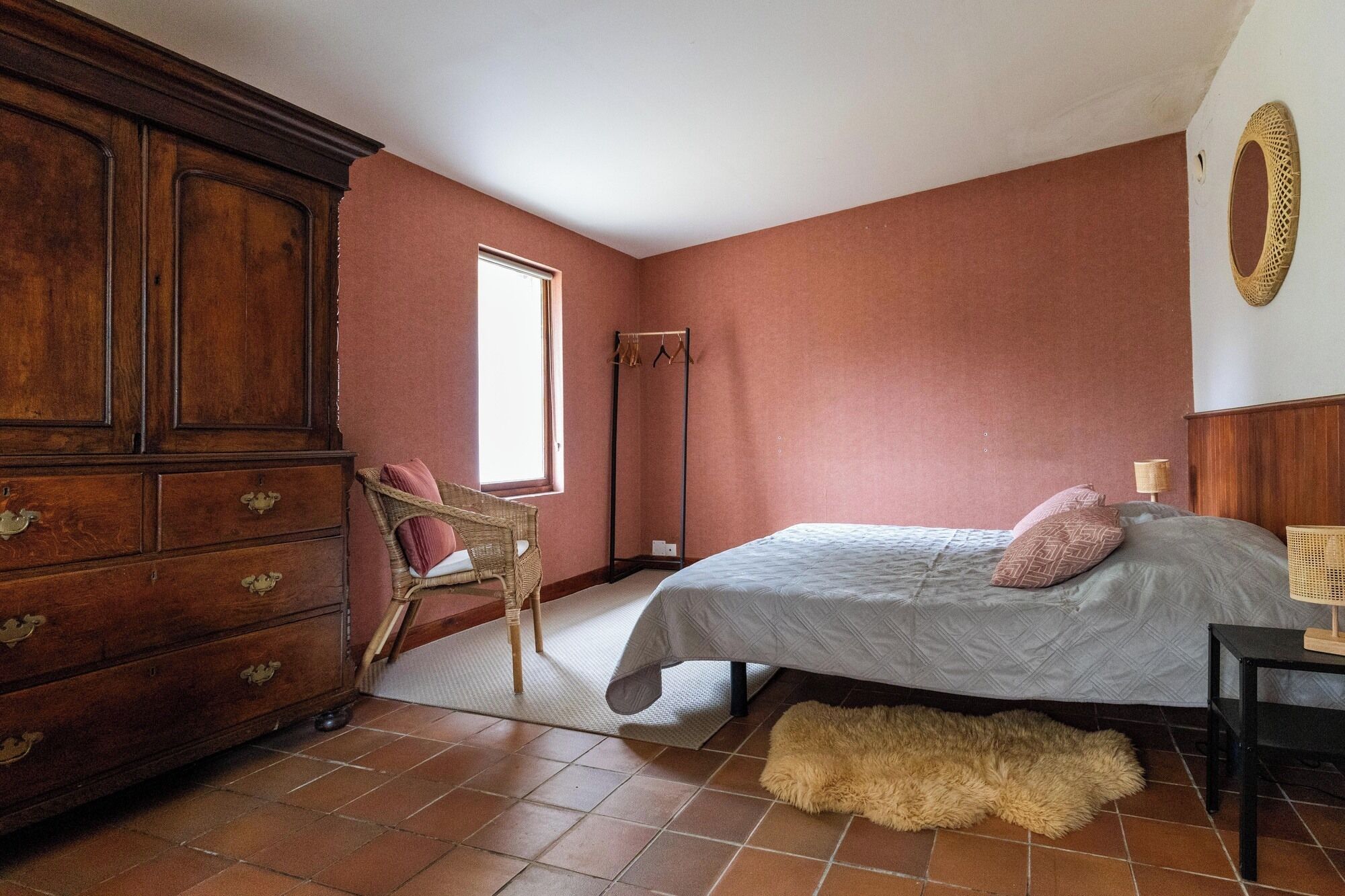 A bedroom with a large wooden dresser, a wicker chair, a bed with a grey quilt, and a fluffy rug on a tiled floor.