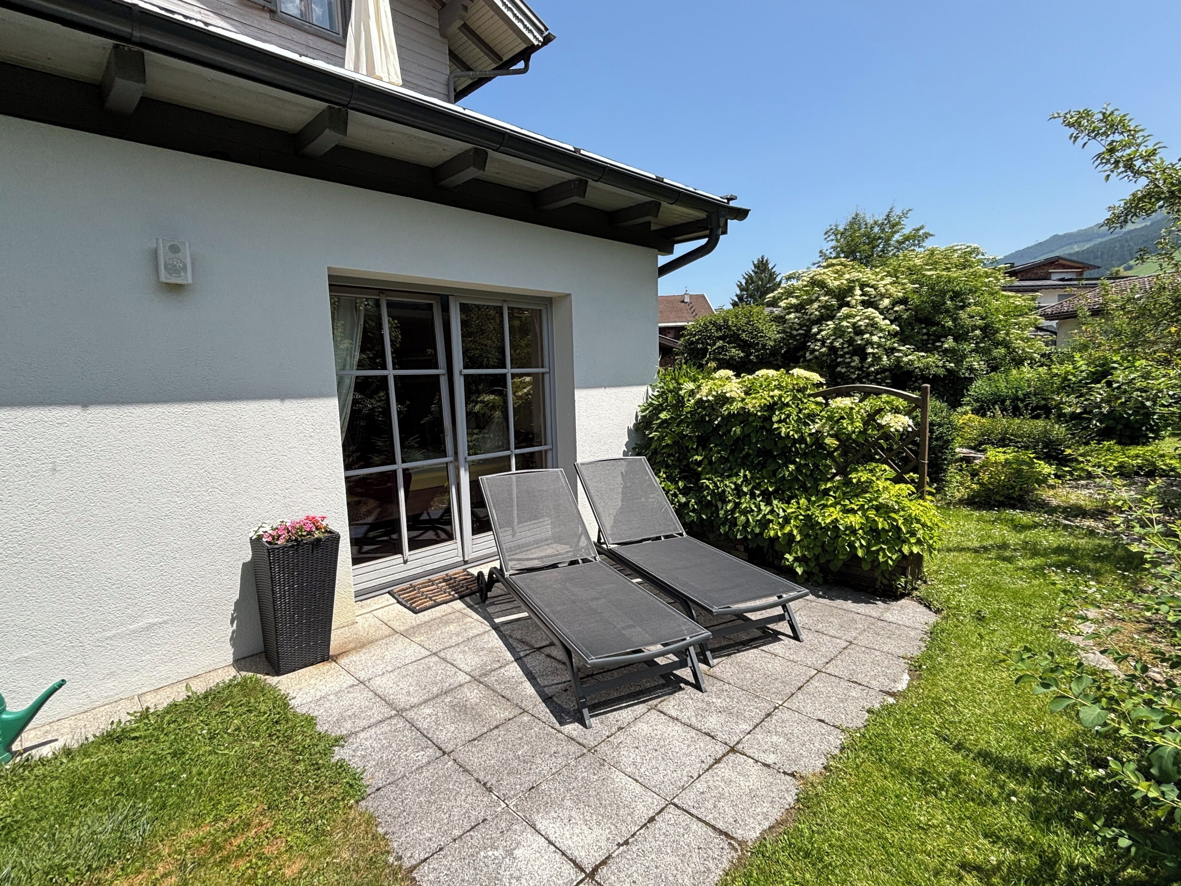 A patio with two lounge chairs and a small table, a white building with a window, and a garden with bushes and trees.