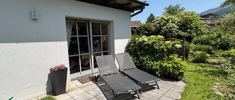 A patio with two lounge chairs and a small table, a white building with a window, and a garden with bushes and trees.