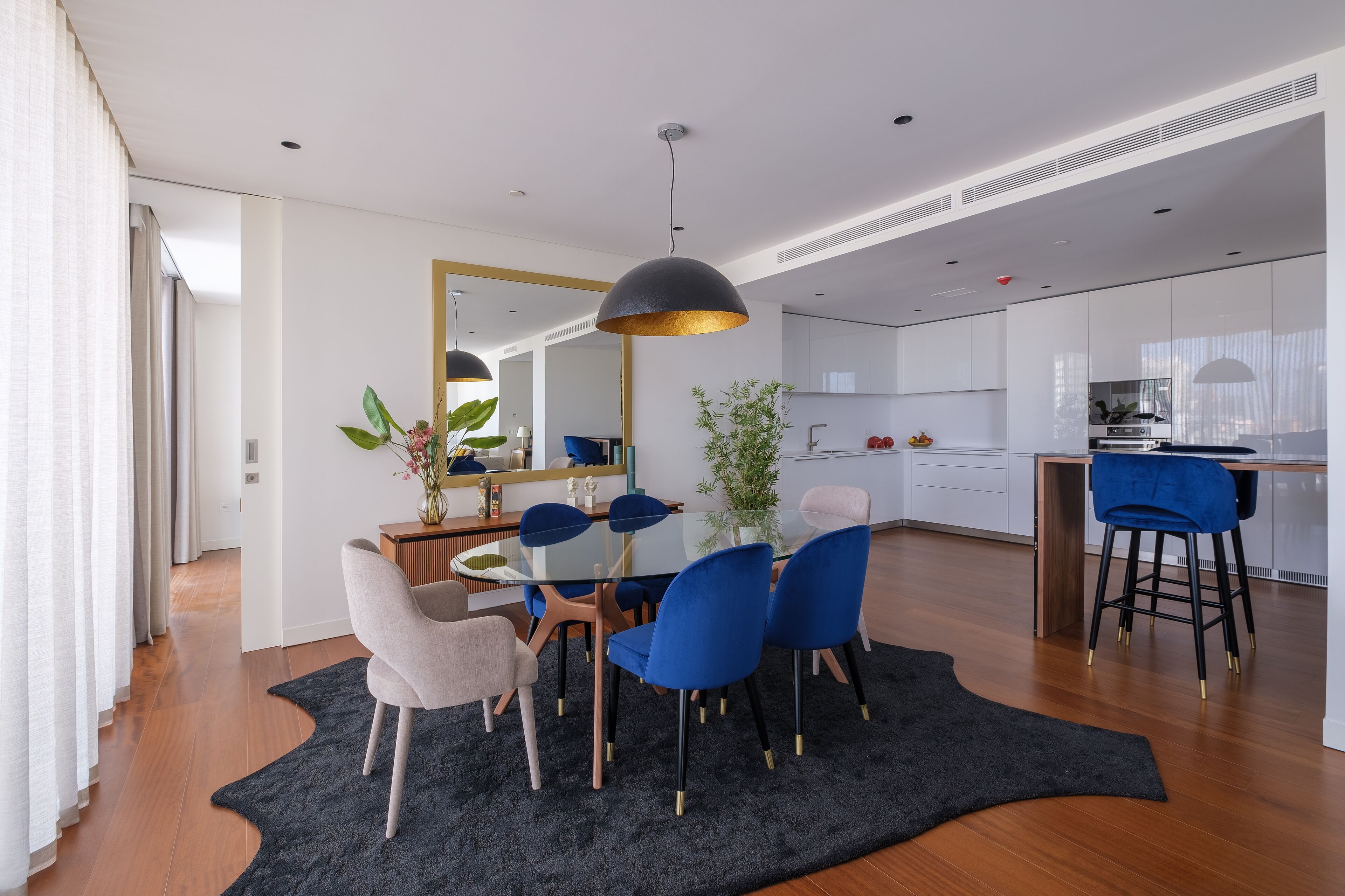 Un comedor moderno con una mesa de vidrio, sillas azules y grises, una lámpara colgante negra y suelo de madera.