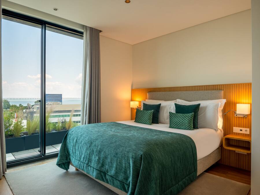 A hotel room with a large bed, two bedside lamps, a wooden headboard, and a view of the city through floor-to-ceiling windows.