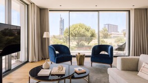 A modern living room with a large TV, two blue armchairs, a beige sofa, and a view of a building and trees through large windows. - Martinhal Lisbon Oriente (Lisbon)