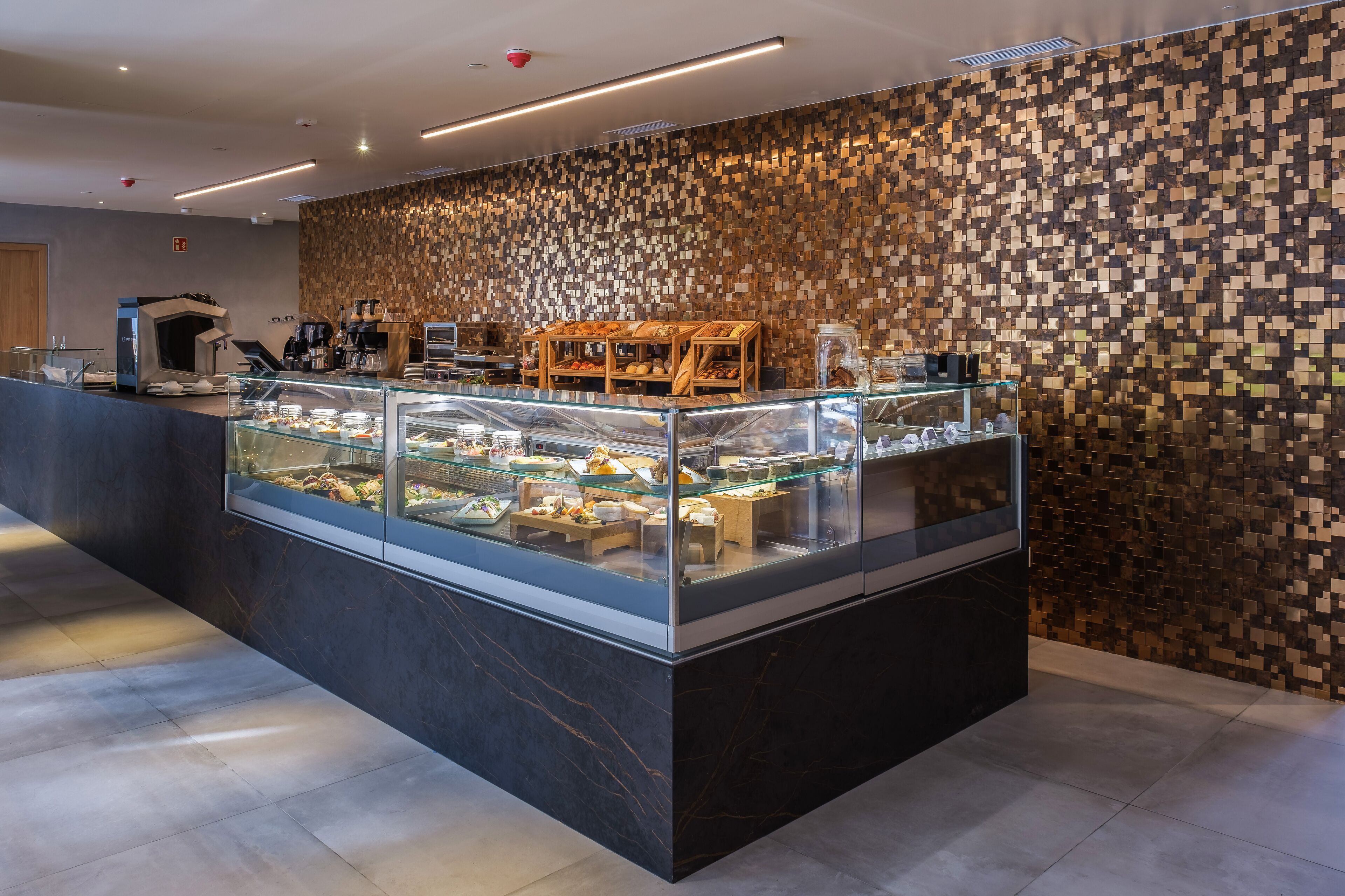 Un bar à petit-déjeuner moderne, avec un mur décoré de mosaïques, une machine à café et une variété d’aliments présentés dans une vitrine.