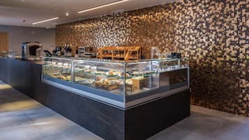 Un bar à petit-déjeuner moderne, avec un mur décoré de mosaïques, une machine à café et une variété d’aliments présentés dans une vitrine.