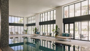 A modern indoor pool area with clear glass walls, lounge chairs, and potted plants. - Martinhal Lisbon Oriente (Lisbon)