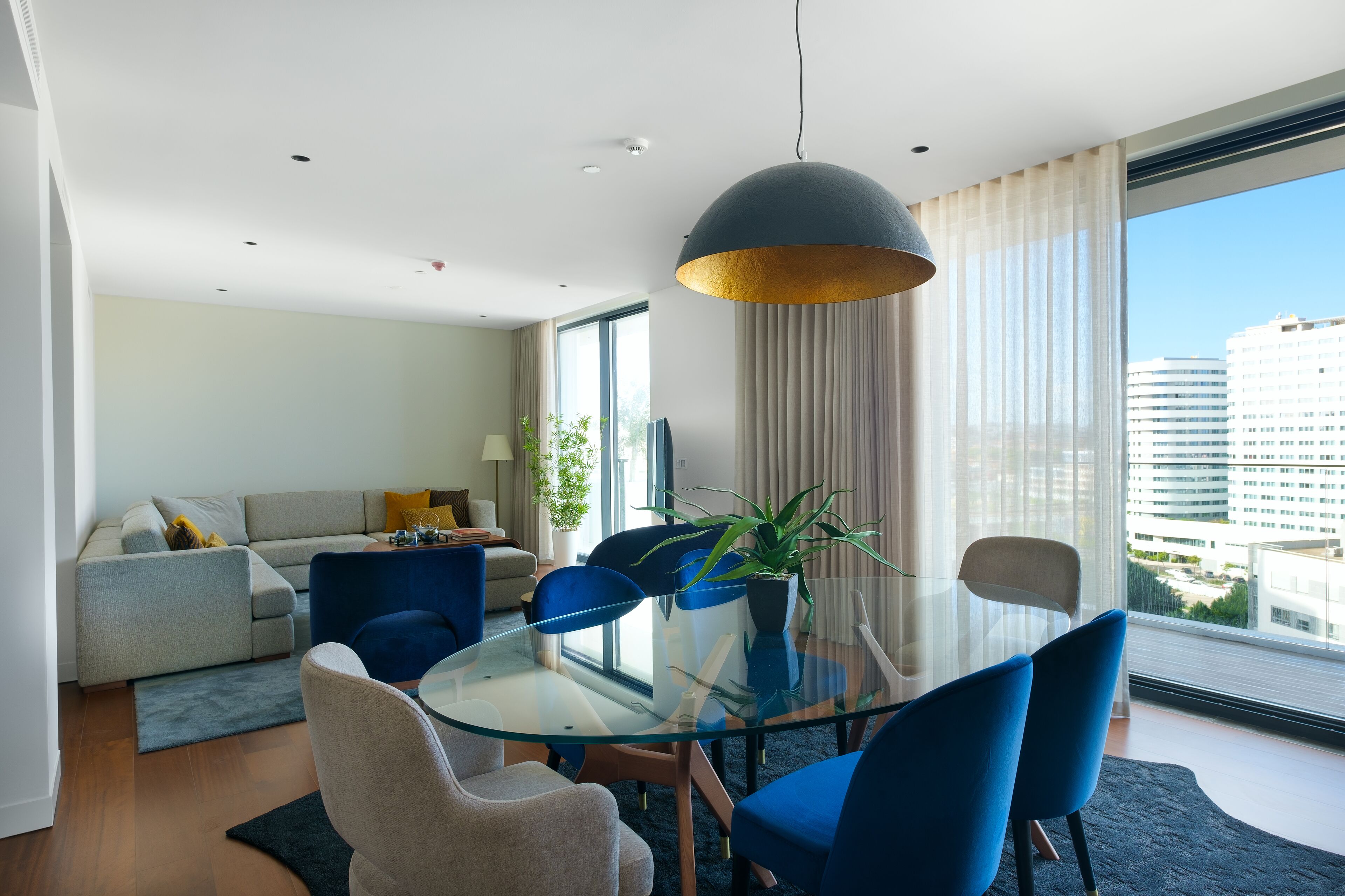A modern living room with a glass dining table, blue chairs, a sofa, and a large window with a city view.