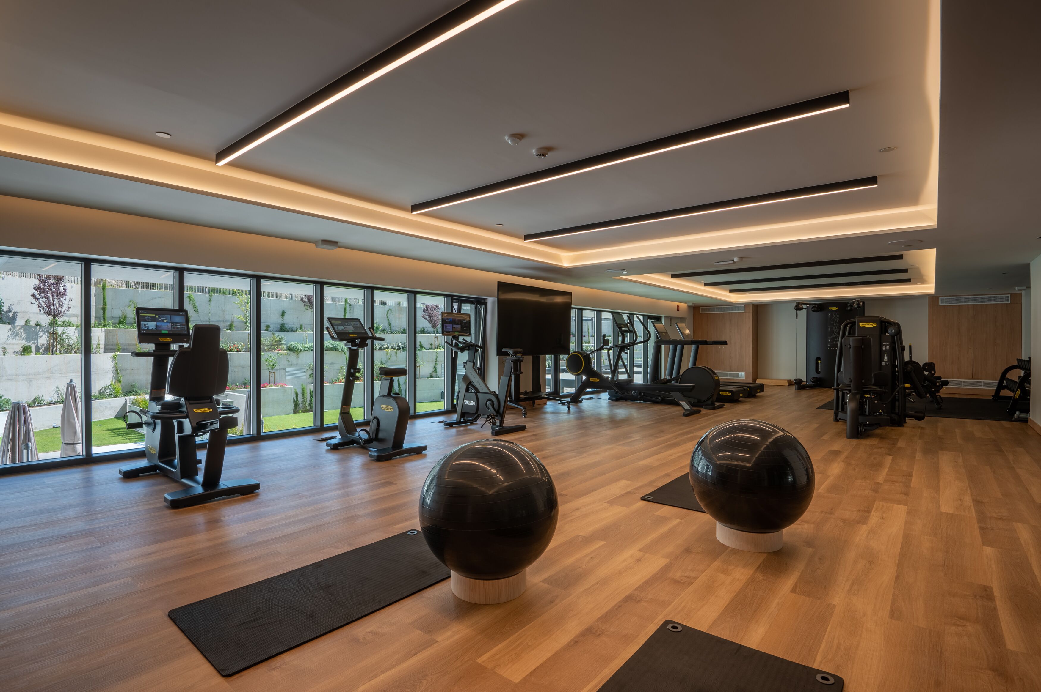 A modern gym with exercise equipment, large windows, and a wooden floor.