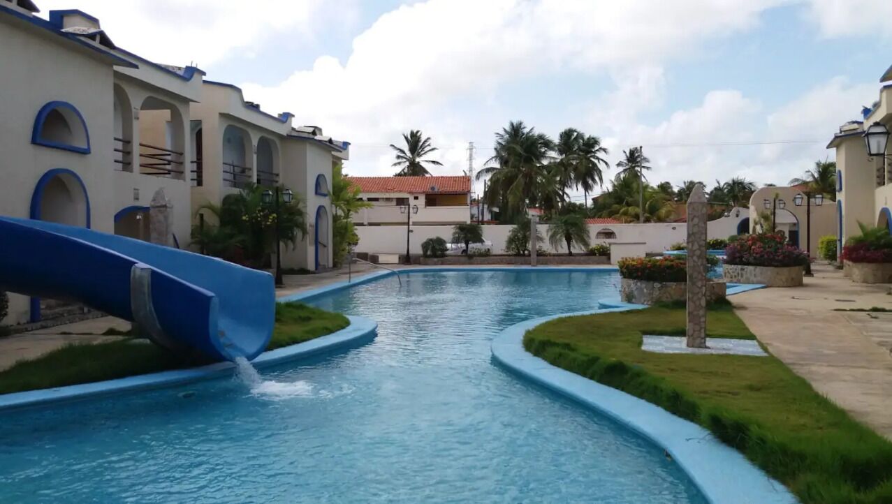 Uma piscina com um escorrega azul, cercada por edifícios com janelas em arco e uma palmeira.