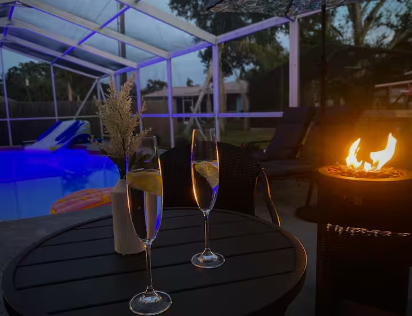 Un espace aménagé au bord de la piscine, avec une table, deux verres contenant une boisson, un vase décoratif et un foyer.