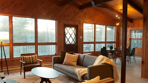 A cozy cabin interior with wooden walls, large windows, a sofa, and a dining area. - Stratton Chalet | Barrel Sauna, Hot Tub & Views (Winhall)