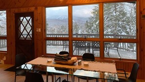 A wooden cabin interior with a dining table, chairs, and a large window offering a snowy mountain view. - Stratton Chalet | Barrel Sauna, Hot Tub & Views (Winhall)