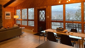 A cozy cabin interior with a wooden ceiling, large windows, a dining area, and a sofa. - Stratton Chalet | Barrel Sauna, Hot Tub & Views (Winhall)