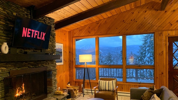 A cozy cabin interior with a stone fireplace, a Netflix sign, a large window with a view of snow-covered trees, and a comfortable seating area. - Stratton Chalet | Barrel Sauna, Hot Tub & Views (Winhall)