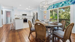 Une cuisine moderne avec des meubles blancs, un coin repas avec des chaises en osier et une vue sur la végétation tropicale grâce à de grandes fenêtres.