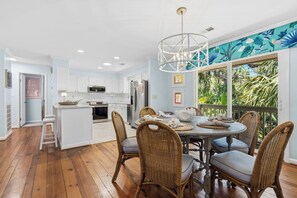 Una cocina moderna con armarios blancos, un comedor con sillas de mimbre y vistas a follaje tropical a través de amplios ventanales.
