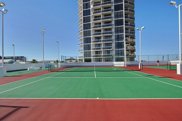 Un court de tennis avec un filet, entouré d’une clôture, et un grand immeuble en arrière-plan.