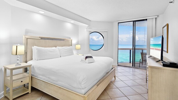 A hotel room with a bed, a nightstand, a TV, and a view of the ocean through a window.