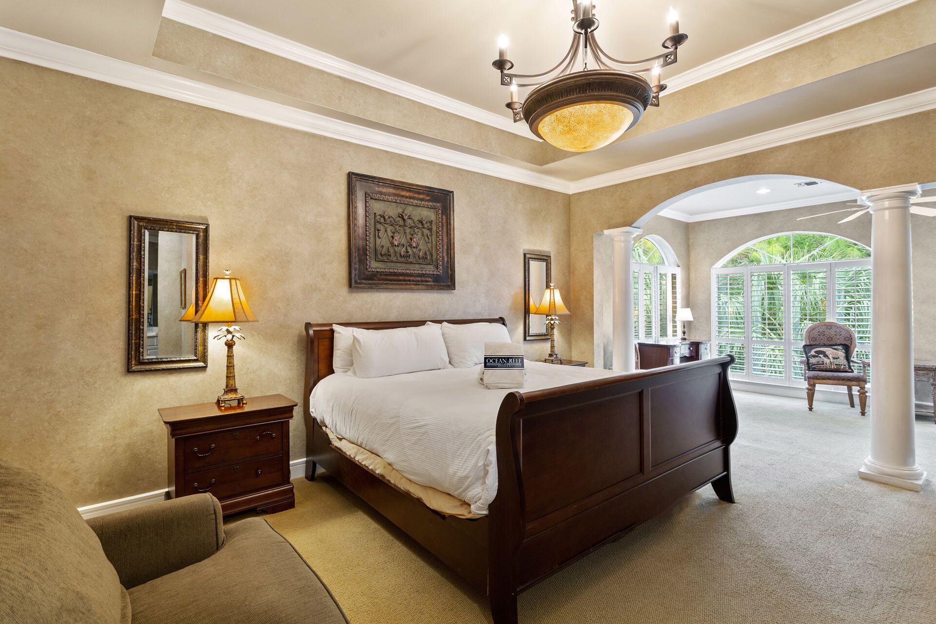 A spacious bedroom with a large bed, two bedside tables with lamps, a wooden nightstand, a chair, and a view of a lush garden through a large window.