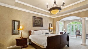 Une chambre spacieuse avec un grand lit, deux tables de chevet avec des lampes, une petite table en bois, une chaise et une vue sur un jardin luxuriant, visible à travers une grande fenêtre.